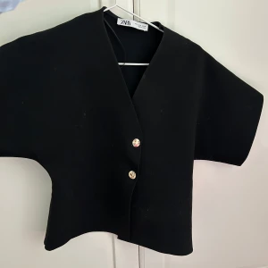 Svart kortärmad stickad från ZARA - Finns ej att köpa längre! Säljer en svart kortärmad jacka från ZARA med två dekorativa knappar framtill. Jackan har en rak och boxig passform och är perfekt för att ge en stilren touch till din outfit.