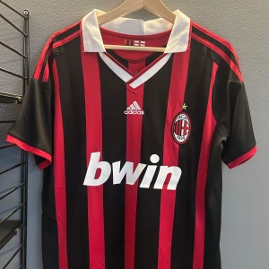 AC Milan fotbollströja Maldini #3 - Säljer en klassisk AC Milan fotbollströja med Maldini och nummer 3 på ryggen. Tröjan är svart och röd med vita detaljer, Adidas-logga och Bwin sponsortryck. Kortärmad modell med klubbmärke på bröstet och randigt mönster. Helt ny med tagg en replika