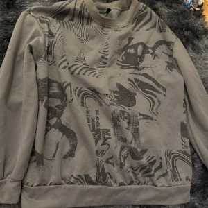 Beige sweatshirt med abstrakt mönster - Säljer en beige sweatshirt med unikt abstrakt mönster i mörkare toner. Tröjan har rund halsringning och långa ärmar. Perfekt för dig som vill sticka ut med en cool och modern stil.