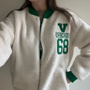 Vit teddyjacka med gröna detaljer  - Vit teddyjacka med gröna muddar och krage. Jackan har dragkedja, två fickor och broderad text i grönt💚