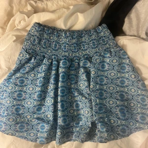 Blå mönstrad kortkjol med mönster - Supersöt blå kortkjol med smock i midjan och ett detaljerat mönster i olika blå nyanser. Kjolen har en luftig passform och är perfekt för varma dagar. Lätt att matcha med en enkel topp eller hoodie.