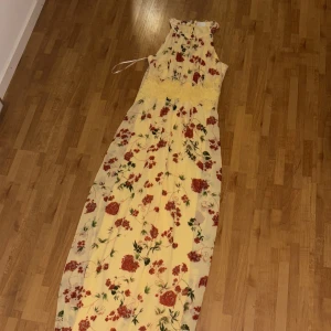 Gul blommig långklänning från Vila - Säljer en lång gul klänning från Vila med rött och grönt blommönster. Klänningen har hög hals, är ärmlös och har spetsdetalj i midjan. Tillverkad i återvunnen polyester. Perfekt för sommarens tillfällen!