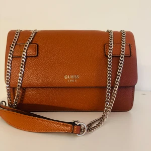 Brun axelväska från Guess - Säljer en snygg brun axelväska från Guess med kedjerem och stilren design. Väskan har en klassisk rektangulär form och är tillverkad i skinnliknande material med struktur. Perfekt för att lyfta din outfit med en trendig accessoar.