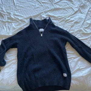 Svart stickad tröja med dragkedja från Jack & Jones - Mörk stickad tröja från Jack & Jones med halv dragkedja framtill. Tröjan har ribbad struktur och hög krage, perfekt för kyligare dagar. Enkel och stilren design som passar till mycket.