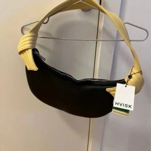 Svart och vit axelväska från HVISK - Cool axelväska från HVISK i svart och vitt med bred, vadderad gul axelrem. Väskan har dragkedja upptill och är tillverkad i veganskt material. Perfekt för att lyfta din outfit med en modern touch. Köpt 2021 men har aldrig kommit till användning 