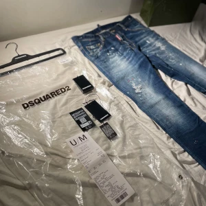 DSQUARED2 jeans med färgstänk - Snygga blå jeans från DSQUARED2 med unika färgstänk och slitningar. Klassisk femficksmodell med orange kontrastsömmar och röd DSQUARED2-logga vid fickan. Perfekta för dig som vill sticka ut med en cool streetstil. KVITTO FINNS