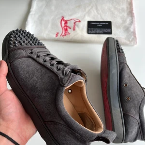 Christian Louboutin skor - Säljer ett par gråa sneakers från Christian Louboutin i mocka med svarta nitar på tån. Skorna har snörning och den klassiska röda sulan. Perfekta för dig som vill sticka ut med en lyxig och edgy stil.