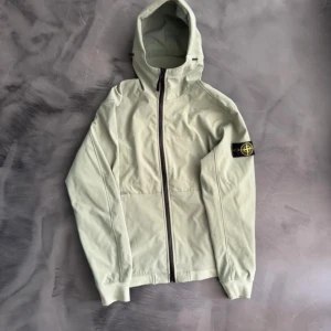 Ljusgrön vindjacka från Stone Island - Snygg ljusgrön vindjacka från Stone Island med huva och hel dragkedja framtill. Jackan har den klassiska logotypen på vänster ärm och två sidofickor. Perfekt för vår och höst när du vill ha en stilren och sportig look.