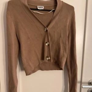 Beige croppad kofta med knappar från Gina Tricot - Säljer en beige croppad kofta från Gina Tricot med långa ärmar och stora dekorativa knappar framtill. Perfekt att slänga över en topp för en chill look. Tunn och mjuk i materialet.