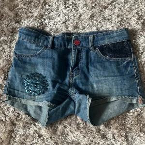 Blå jeansshorts med paljettdetaljer från Desigual - Snygga blå jeansshorts från Desigual med paljetter på ena fickan fram och ett paljettband bak. Shortsens knapp är röd och de har klassisk femficksdesign. Perfekta för dig som gillar unika detaljer och denim.Shortsen är i barnstolek.