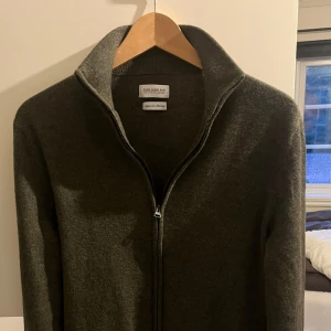 merinoullskofta från These Glory Days - Säljer en stilren olivgrön kofta från These Glory Days i mjuk superior merinoull. Koftan har dragkedja framtill, hög krage och lång ärm. Perfekt för lager-på-lager och passar till många olika outfits. Nypris:1099. 