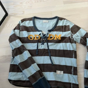 Randig långärmad tröja från Odd Molly - Säljer en långärmad tröja från Odd Molly med breda bruna och ljusblå ränder. Tröjan har snörning framtill vid halsen och broderad logga i gult. Mjuk bomullskänsla och avslappnad passform.