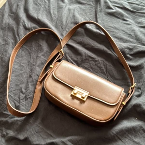 Brun axelväska med gulddetaljer - Säljer en stilren brun axelväska med justerbart axelband och snyggt guldfärgat spänne framtill. Väskan har en klassisk rektangulär form och är perfekt för att bära det viktigaste. Enkel och tidlös design som passar till många outfits.