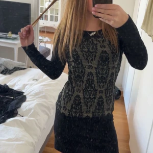 Svart och beige mönstrad klänning från Yest - Kortklänning från Yest i svart med dekorativt mönster framtill. Klänningen har lång ärm och rund halsringning. Materialet känns mjukt och stretchigt, perfekt för dig som gillar en tight passform.