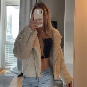 Krämvit teddyjacka med dragkedja - Säljer en krämvit teddyjacka med silvriga dragkedjor. Jackan är croppad och har en oversized passform. Mjuk och fluffig känsla som ger en trendig look!