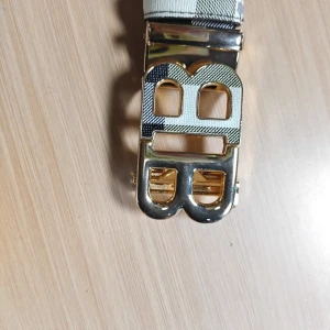 Burberry bälte - Säljer ett trendigt bälte med rutigt mönster i beige, svart, vitt och grått. Bältet har ett stort, guldfärgat BB-spänne framtill som ger en lyxig känsla. Perfekt accessoar för att lyfta din outfit.