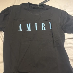 Svart t-shirt från Amiri med blå logga - Snygg svart t-shirt från Amiri med stor blå logotyp på bröstet. Klassisk rund halsringning och korta ärmar. Tillverkad i 100% bomull för en skön och mjuk känsla. Perfekt för en stilren och trendig look.