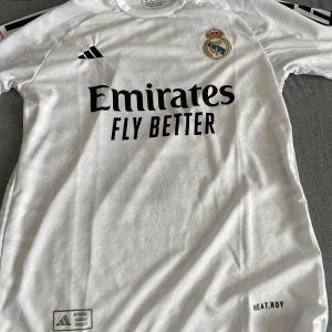 Real Madrid fotbollströja Mbappé 9 - Säljer en vit Real Madrid fotbollströja från Adidas med Mbappé och nummer 9 på ryggen. Tröjan har korta ärmar, klubbmärke på bröstet och sponsortryck framtill. Perfekt för fans som vill visa sitt stöd på matchdagar.