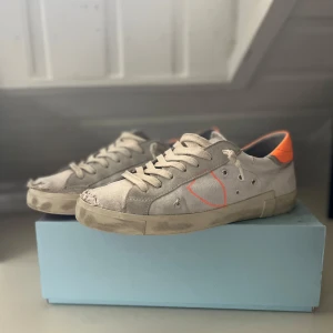 Beige sneakers från Philippe Model med orange detalj - Säljer ett par beige sneakers från Philippe Model med orange häl och sula. Skorna har snörning, rund tå och är tillverkade i mocka och syntet. Perfekta för dig som gillar stilrena men ändå unika sneakers.