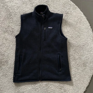 Mörkblå fleeceväst från Patagonia - Snygg mörkblå fleeceväst från Patagonia med dragkedja framtill och tre fickor med dragkedja. Klassisk ståkrage och diskret logga på bröstet. Perfekt lager för kyliga dagar.
