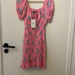 Rosa klänning från bikbok  - Supersöt rosa klänning med blått och grönt blommönster från Bik Bok. Klänningen har puffärmar, smock över byst och midja samt volang nertill. Perfekt för sommarens alla tillfällen!