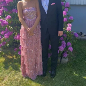 Balklänning  - Säljer en superfin rosa långklänning med glittriga paljetter och pärlor upptill. Klänningen är axelbandslös och har volangdetaljer på kjolen. Perfekt för bal eller festliga tillfällen!