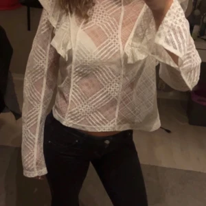 Vit blus med volangdetaljer - Säljer en vit blus i transparent material med rutigt mönster och volangdetaljer vid axlarna. Blusen har långa ärmar. Fin till fest