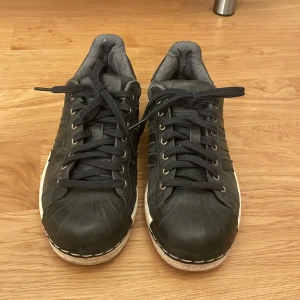 Svarta Adidas sneakers med vita sulor - Säljer ett par svarta Adidas sneakers med klassiska tre ränder på sidan och vit sula. Skorna har snörning och rund tå, samt diskret Adidas-logga där fram. 