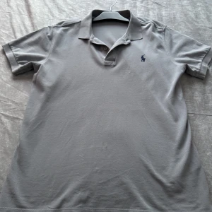 Grå pikétröja från Ralph Lauren - Säljer en klassisk grå pikétröja från Ralph Lauren med broderad logga på bröstet. Tröjan har korta ärmar, krage och knappar vid halsen. Perfekt för en stilren och avslappnad look.
