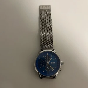 Silverfärgad klocka från BOSS - Stilren klocka från BOSS med blå urtavla och tre mindre sub-dials. Armbandet är i silverfärgat mesh-stål och har en klassisk spänne. Perfekt för den som gillar en elegant och modern look. Oanvänd så 10/10 skick.