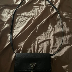 Svart axelväska från Guess - Säljer en stilren svart axelväska från Guess med guldfärgad logga framtill. Väskan har lång axelrem, flera fack inuti och stängs med tryckknapp. Perfekt för att bära det viktigaste med stil.
