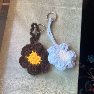Virkat blommigt nyckelring-set - Två handgjorda virkade nyckelringar i form av blommor. En är brun med gul mitt och den andra är ljusblå med vit mitt. Perfekt som söt accessoar till väskan eller nycklarna. Ger riktig sommar vibes 