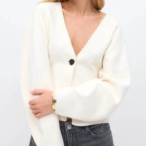 Vit kofta - Säljer en stilren vit kofta från Gina med djup v-ringning och en dekorativ guldig knapp framtill. Koftan har långa ärmar och en rak, något croppad passform. Perfekt att styla med jeans eller kjol. Lappen kvar💕