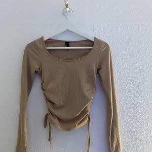 Beige ribbad topp med dragsko från SHEIN - Trendig beige långärmad topp från SHEIN med ribbad struktur och dragsko i sidan som ger en snygg rynkad effekt. Perfekt att styla med jeans eller kjol för en cool look.