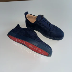 Mörkblå sneakers från Christian Louboutin - Säljer ett par mörkblå sneakers från Christian Louboutin i mocka med klassisk röd sula och snörning. Skorna har rund tå och platt sula, perfekt för dig som vill sticka ut med exklusiva detaljer.