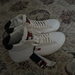  Tommy sneakers - Säljer ett par fräscha vita höga sneakers från Tommy Jeans med klassisk logga på sidan och plös. Skorna har vita snören och detaljer i svart och rött. Perfekta för dig som gillar stilrena skor. De är dessutom oanvända och helt nya