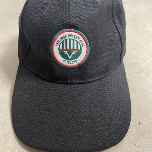 Svart keps Frölunda Hockey Club Göteborg - Svart keps med broderad Frölunda Hockey Club-logga framtill och texten 'Göteborg' bak. Justerbar rem baktill. Klassisk form och böjd skärm. Perfekt för hockeyfans!