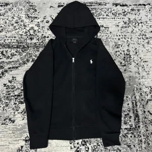 Svart hoodie från Polo Ralph Lauren - Klassisk svart hoodie från Polo Ralph Lauren med dragkedja och huva. Tröjan har det ikoniska broderade logotypen på bröstet och är tillverkad i mjukt material. Perfekt för en avslappnad och stilren look.