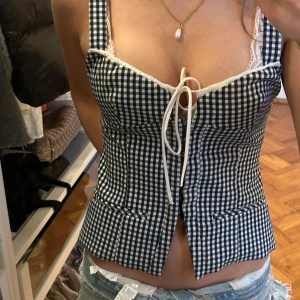 Svartvit rutig korsettopp med knytning från zara! Säljs inte längre - Supersöt korsettopp i svartvitt rutigt mönster med knytning framtill och spetsdetaljer vid bysten. Toppen har smala axelband och figurnära passform, perfekt för att lyfta vilken outfit som helst.