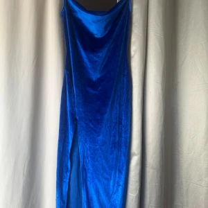 Blå långklänning i sammet med slits - Säljer en snygg blå långklänning i sammetsmaterial med smala axelband och en elegant slits framtill. Klänningen har en draperad urringning och sitter figurnära. Perfekt för dig som vill sticka ut med en färgstark look.