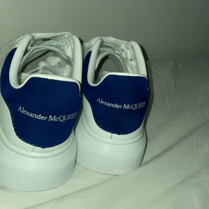 Vita sneakers från Alexander McQueen med blå häl - Säljer ett par vita sneakers från Alexander McQueen med blå mockadetalj på hälen och vit snörning. Skorna har en chunky sula och klassisk rund tå. Perfekta för dig som gillar stilrena och trendiga sneakers.