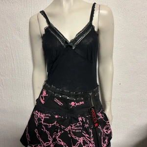 Svart och rosa kjol från Poizen Industries - Cool svart minikjol med rosa kedjemönster och detaljer från Poizen Industries. Kjolen har dragkedja framtill, säkerhetsnålar i midjan och spetskant nedtill. Märket syns tydligt på etiketten och trycket.