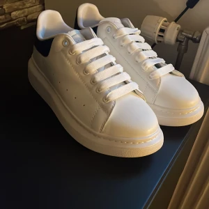 Vita sneakers från Alexander McQueen med blå häl - Säljer ett par vita sneakers från Alexander McQueen med blå detalj på hälen och klassisk snörning. Skorna har en chunky sula och stilren design, perfekta för dig som gillar exklusiva och trendiga sneakers.