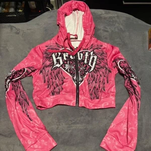 Rosa croppad hoodie med vingar och tryck - Säljer en rosa croppad hoodie med dragkedja från Sekai. Jackan har ett stort vingmotiv och texten 'Beauty' i vitt och svart på bröstet samt detaljerade tryck på ärmarna. Huvan är fodrad i vitt och jackan har långa ärmar.