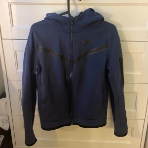 Blå zip-hoodie från Nike - Säljer en blå hoodie från Nike med dragkedja och huva. Jackan har svarta detaljer och Nike-logga på bröstet. Perfekt för träning eller chill. Den är använd några fåtal gånger och har därmed lite knottrigt tyg.