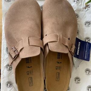 Beige Birkenstock Boston i mocka - Birkenstock Boston – strl 43, färg Taupe – i princip nya!  Säljer ett par nästan helt nya Birkenstock Boston i färgen Taupe, storlek 43 EU. Använda max 5 gånger inomhus – lapparna sitter fortfarande kvar! Säljes då de inte riktigt passar min stil och det blev för dyrt att returnera.  Inköpta för ca 1900 kr. Kan eventuellt ordna köpbevis vid behov, men skicket talar för sig själv. Modellen är från herravdelningen, men funkar lika bra för alla