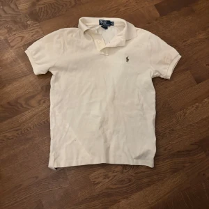 Vit pikétröja från Polo Ralph Lauren - Klassisk vit pikétröja från Polo Ralph Lauren med broderad logga på bröstet. Tröjan har korta ärmar, krage och knappar vid halsen. Perfekt för en stilren och tidlös look. Säljer på grund av att jag har en annan  piké i den här färgen. Och samma storlek. Fick av brorsan😁Pris kan diskuteras