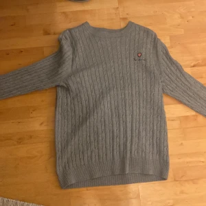 Ljusgrå stickad tröja från Bondeli - Säljer en ljusgrå stickad tröja från Bondeli med klassiskt kabelstickat mönster och rund halsringning. Tröjan har en liten broderad Bondeli-logga på bröstet och ribbade muddar vid ärmslut och nederkant. Perfekt för lager-på-lager-stil.