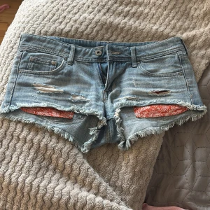 Ljusblå lågmidjade jeansshorts med fransar och paisleydetaljer - Säljer ett par jättesnygga ljusblå jeansshorts med slitna detaljer, fransig kant och röda paisleymönstrade tygdetaljer i fickorna. de är i storlek S och jag säljer pågrund av att de tyvärr inte passar på mig längre. Klassisk femficksmodell med knapp och dragkedja. Perfekta för sommaren!🥰🥰🥰 Skriv gärna
