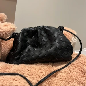 Svart axelväska i flätat skinn - Säljer en svart axelväska i flätat skinn med justerbar rem. Väskan har en mjuk, puffig form och stängs med dragsko. Perfekt för att bära det viktigaste och ger en trendig touch till din outfit.